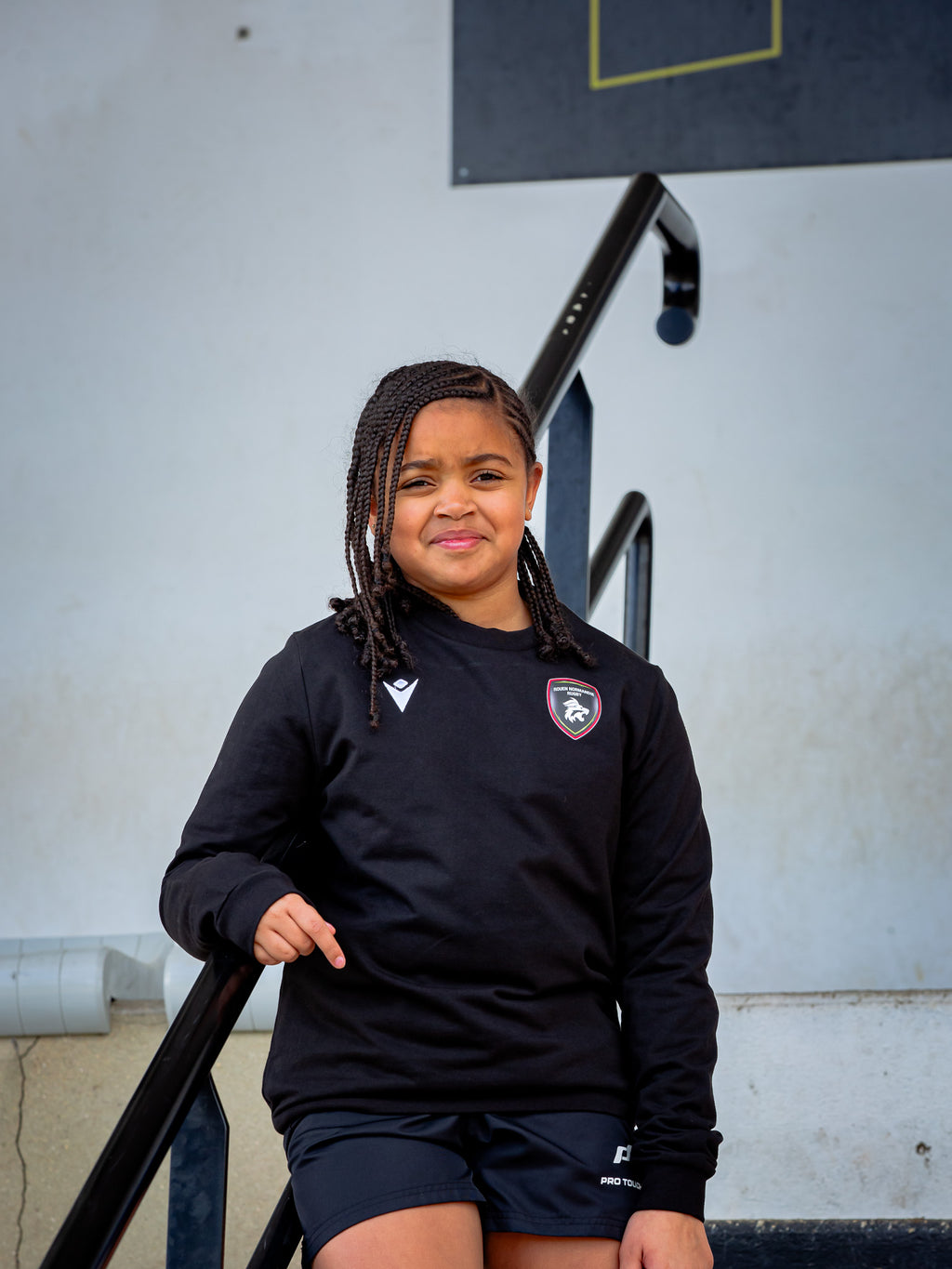 Une jeune fille aux cheveux tressés, vêtue d'un sweat-shirt Rouen Normandie Rugby Noir Axima JR et d'un short sportif, se tient debout, légèrement souriante, sur un escalier extérieur.