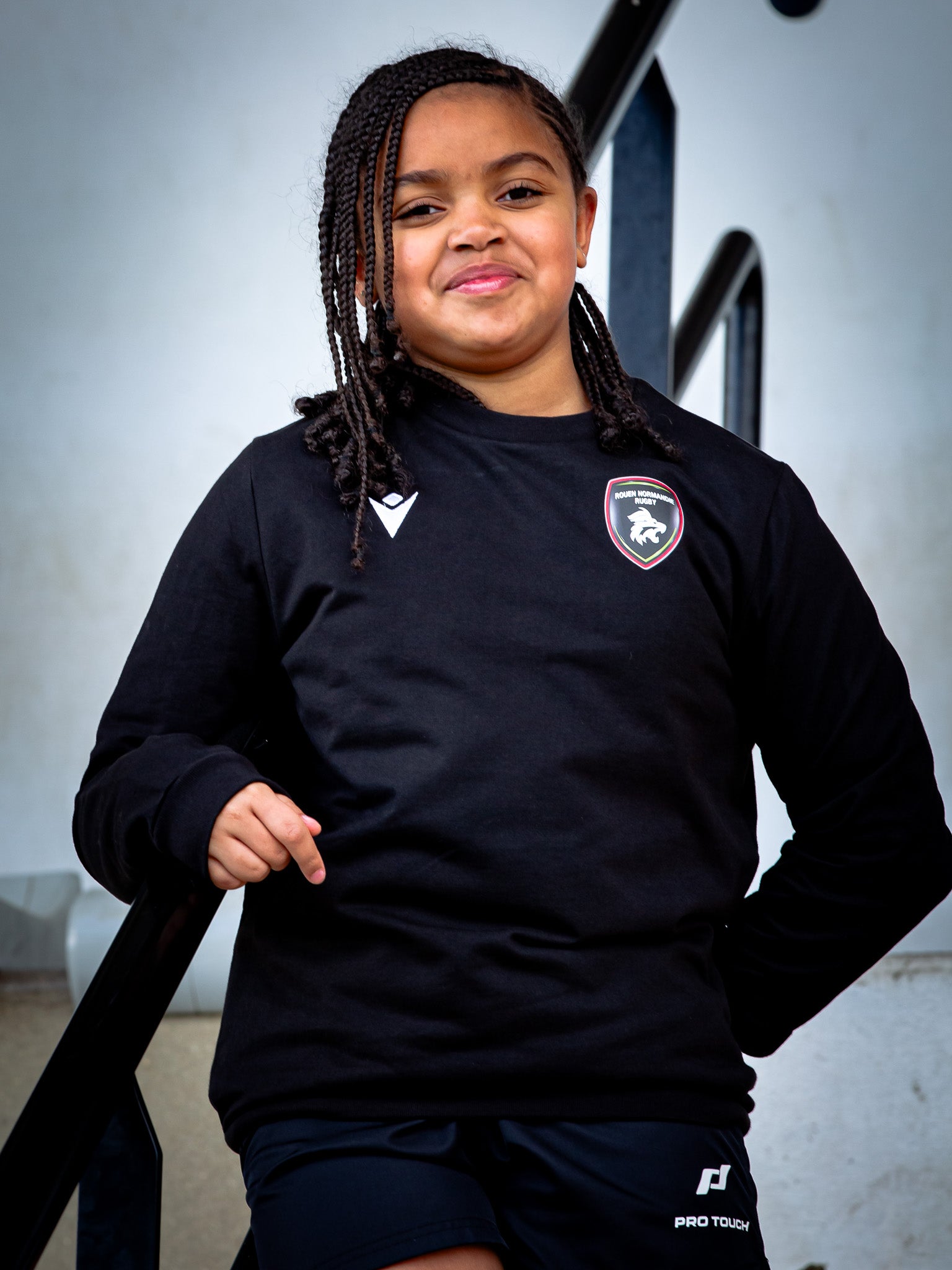 Une jeune fille aux cheveux tressés, vêtue d'un sweat-shirt Rouen Normandie Rugby Noir Axima JR et d'un short, se tient debout sur des marches et sourit avec assurance à la caméra.