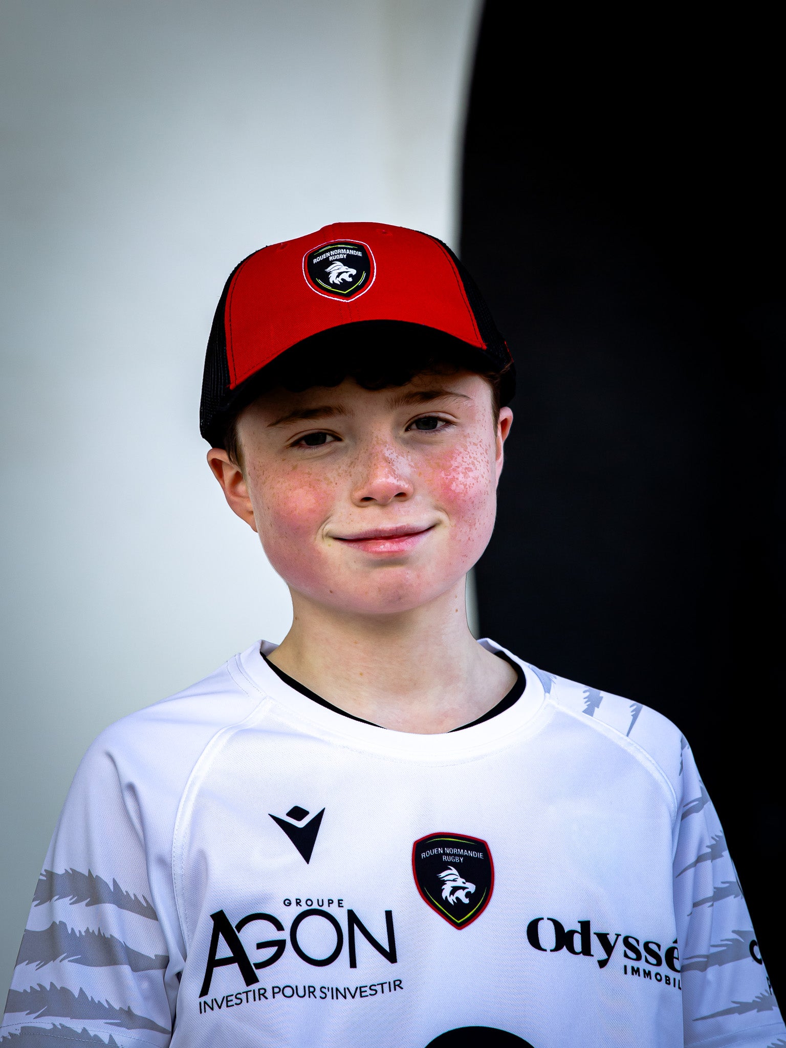 Un jeune garçon vêtu d'une Casquette trucker RNR de Rouen Normandie Rugby et d'un maillot de sport blanc avec logos se tient devant une toile de fond noire et blanche, souriant à l'appareil photo - son style de supporter capture l'esprit Rouen Normandie Rugby.