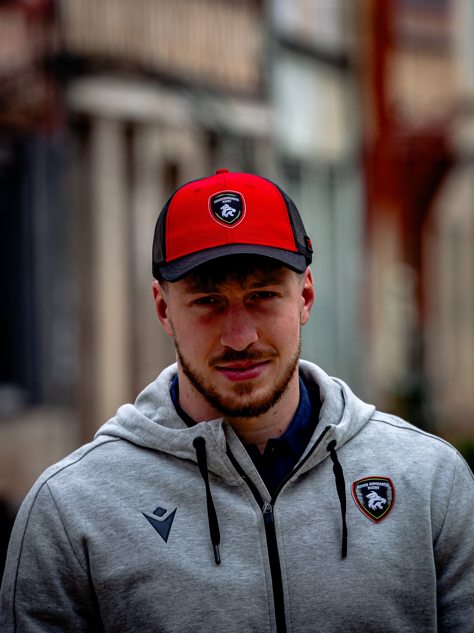 Un homme vêtu d'un camion Casquette RNR de Rouen Normandie Rugby et d'une veste zippée gris clair avec les logos des lions assortis se tient à l'extérieur, la vie urbaine s'estompant derrière lui - le style idéal pour tout vrai supporter de Rouen Normandie Rugby.