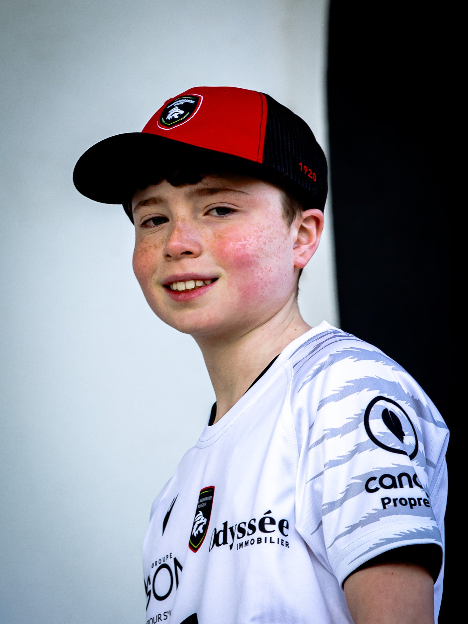 Un jeune garçon à la peau claire et aux taches de rousseur, vêtu d'un camion Casquette RNR et d'un maillot blanc du Rouen Normandie Rugby, se tient devant un fond noir et blanc, souriant doucement à l'appareil photo.