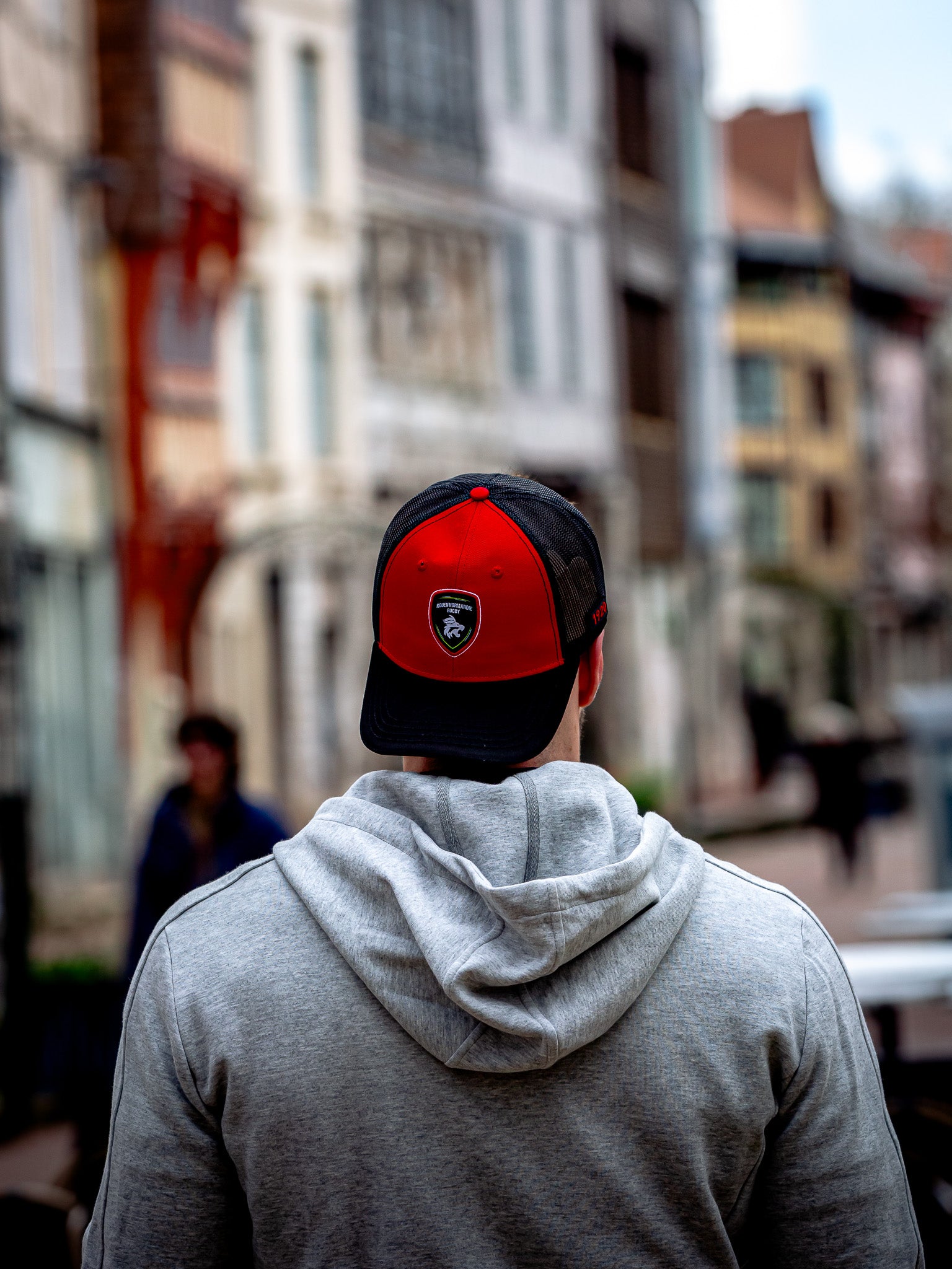 Une personne vêtue d'un sweat à capuche gris et d'un camion Rouen Normandie Rugby Casquette RNR est vue de dos, marchant dans une rue vibrante et floue bordée d'immeubles.