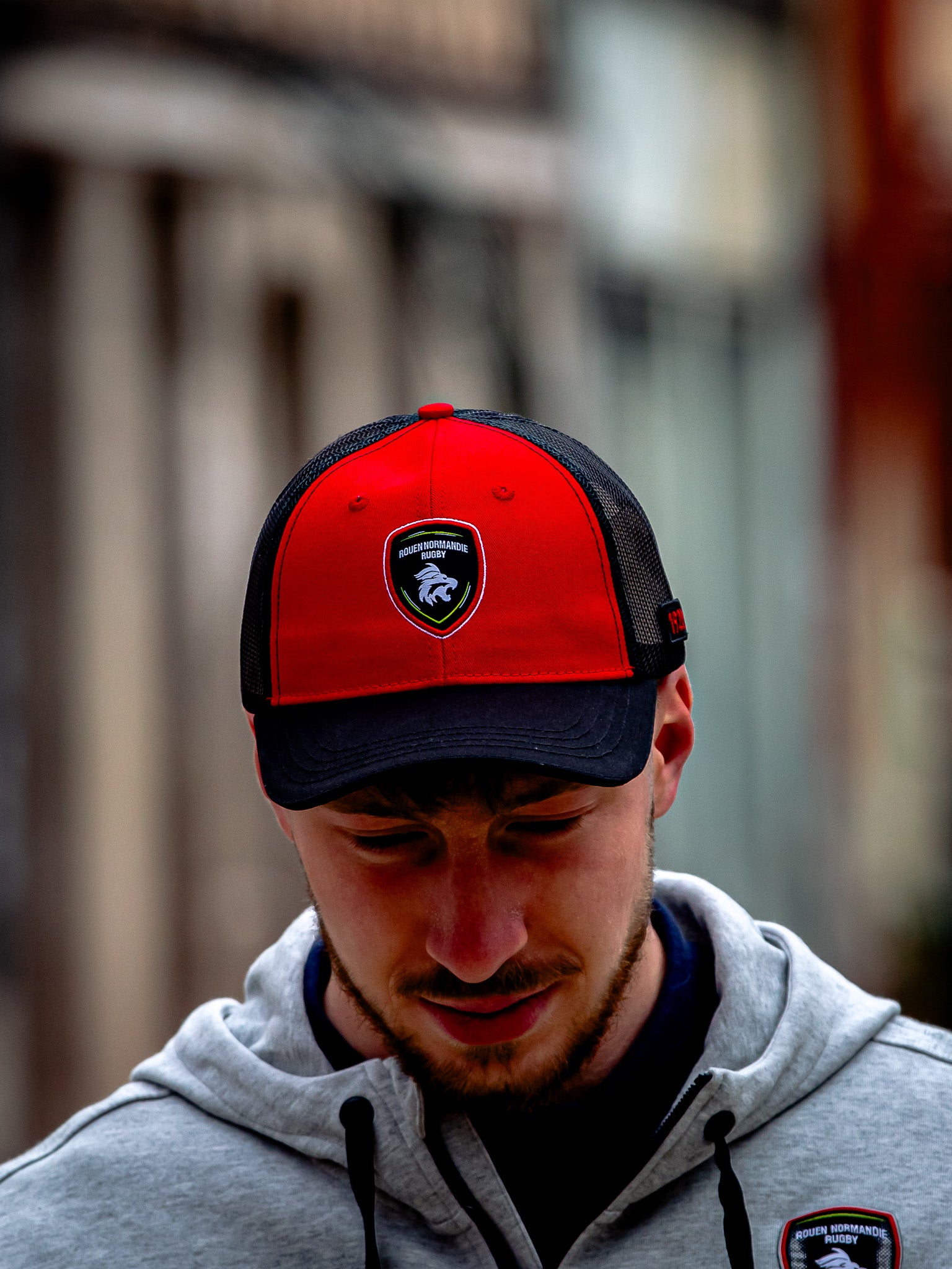 Un homme portant un camion Casquette RNR de Rouen Normandie Rugby et un sweat à capuche gris clair avec l'emblème de l'équipe se tient à l'extérieur, regardant vers le bas. L'arrière-plan est flou.