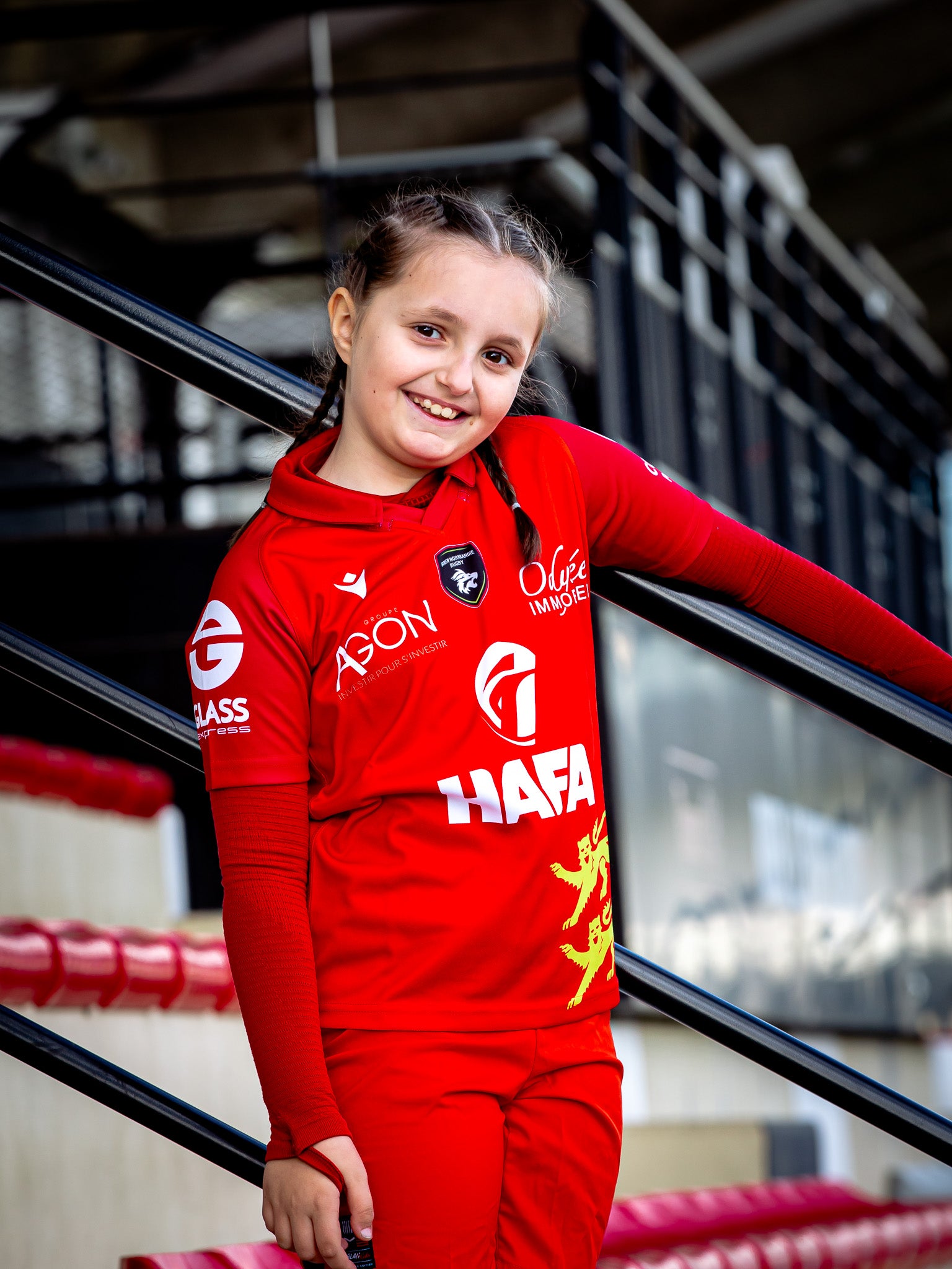Une jeune fille souriante aux cheveux tressés se tient debout sur des escaliers, tenant la rampe et portant le Rouen Normandie Rugby Maillot Exterieur 25-26 Junior avec les logos des sponsors et l'emblème du lion. Les gradins du stade sont visibles à l'arrière-plan.