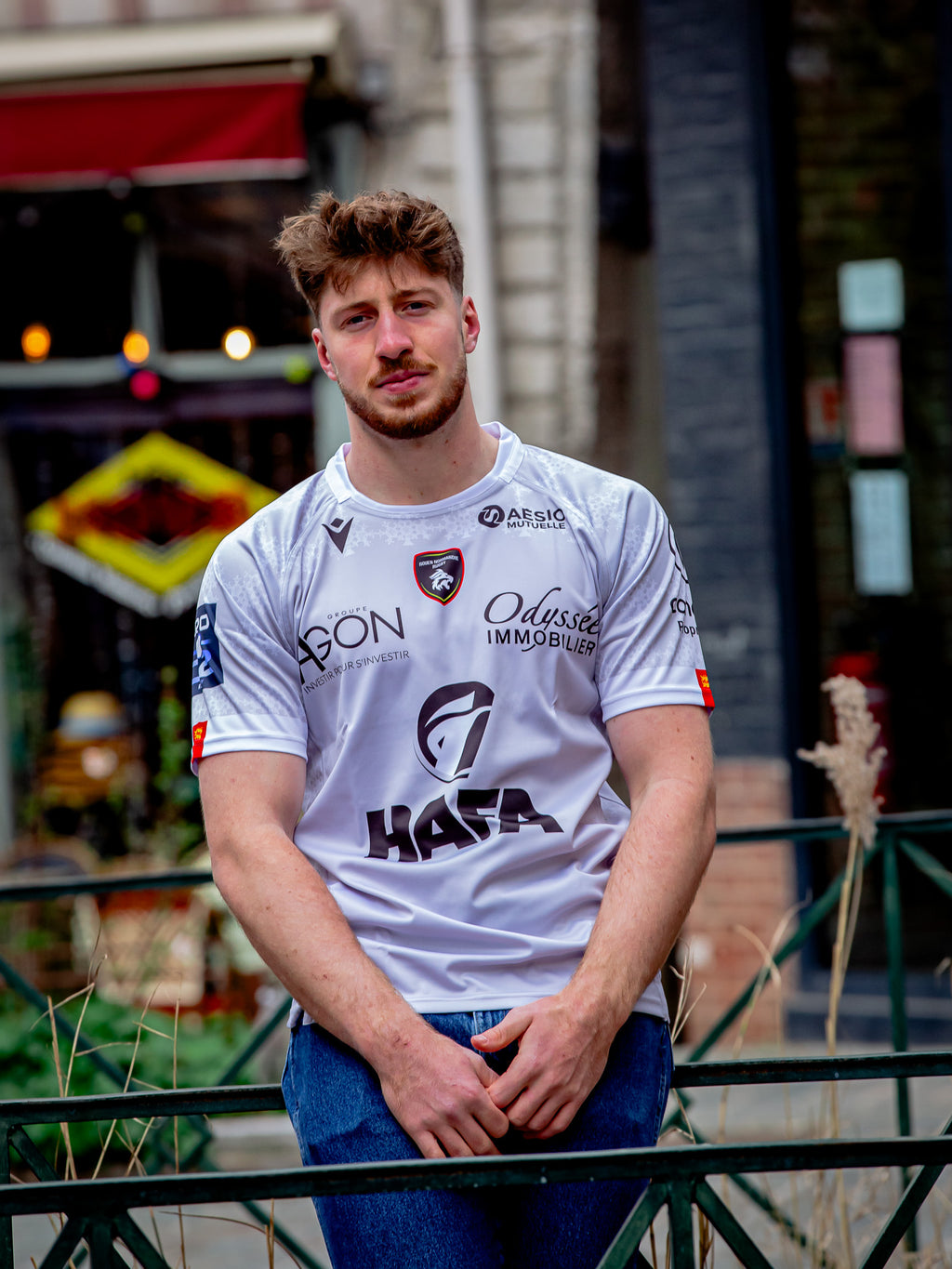 Un jeune homme aux cheveux bruns courts et à la barbe se tient en plein air dans le Rouen Normandie Rugby Maillot Extérieur 23-24 Adulte, vêtu d'un jean bleu, les mains appuyées sur une rambarde métallique. Des bâtiments urbains et de la verdure sont visibles en arrière-plan.