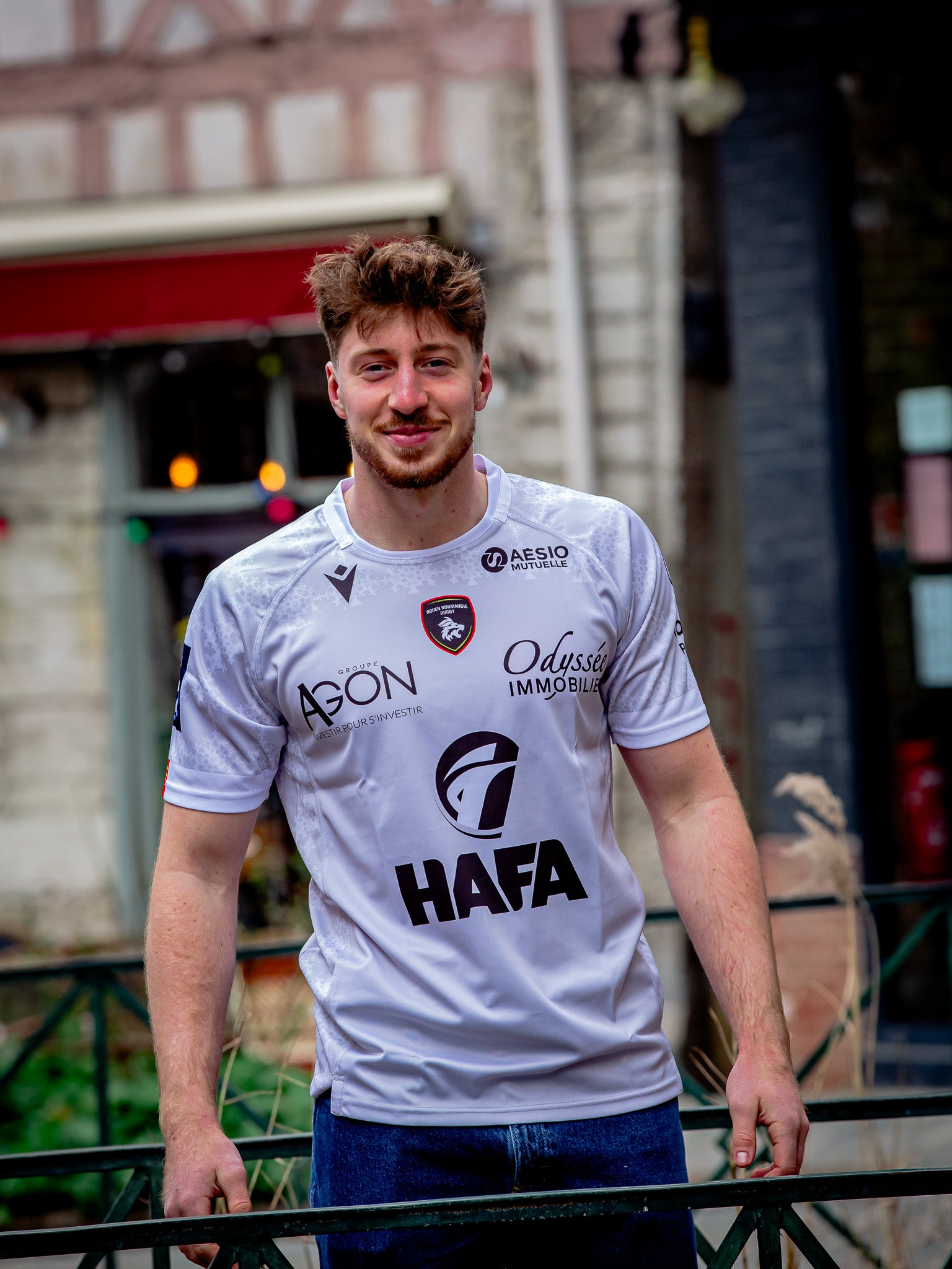 Un jeune homme aux cheveux bruns courts et à la barbe sourit à la caméra en plein air, portant le Rouen Normandie Rugby Maillot Extérieur 23-24 Adulte avec les logos des sponsors et un jean bleu, debout près d'une rambarde métallique et de bâtiments colorés.