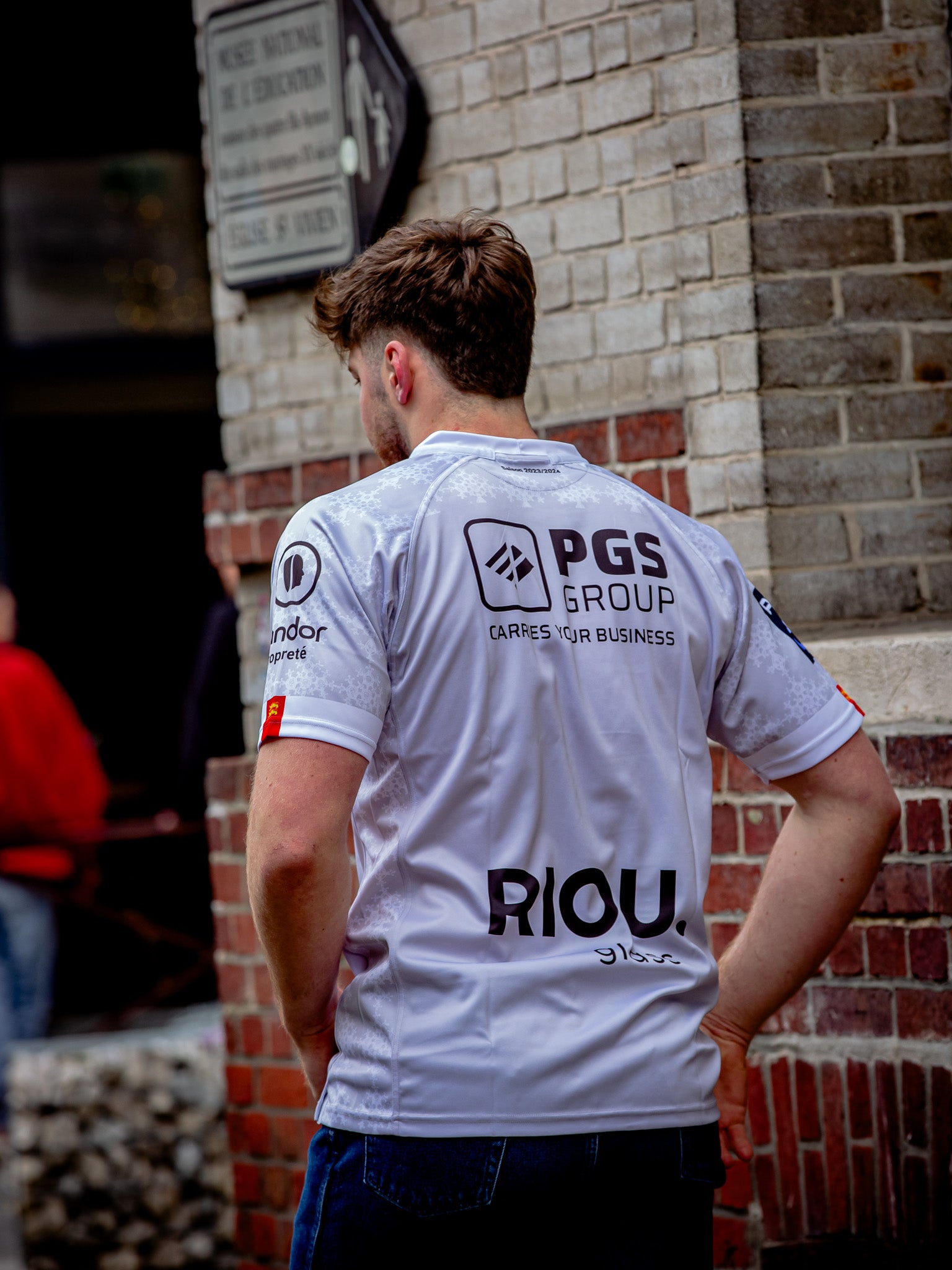 Un homme se tient à l'extérieur, près d'un mur de briques et d'une plaque de rue. Il porte le Rouen Normandie Rugby Maillot Extérieur 23-24 Adulte, une réplique de maillot avec des logos de sponsors tels que PGS GROUP et RIOU visibles dans le dos.