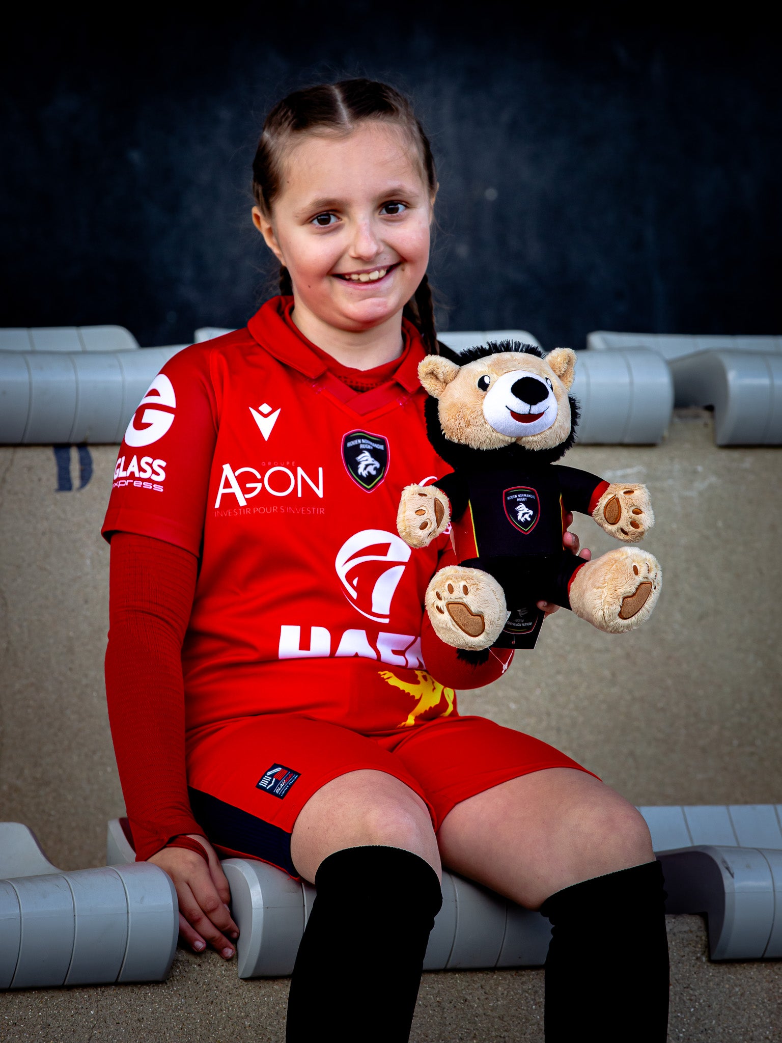 Une jeune fille souriante en tenue de football rouge est assise sur des gradins et tient la Peluche Officielle Rouen Normandie Rugby - Lion Mascotte by Rouen Normandie Rugby, toutes deux portant des maillots assortis, avec un arrière-plan sombre derrière elles.