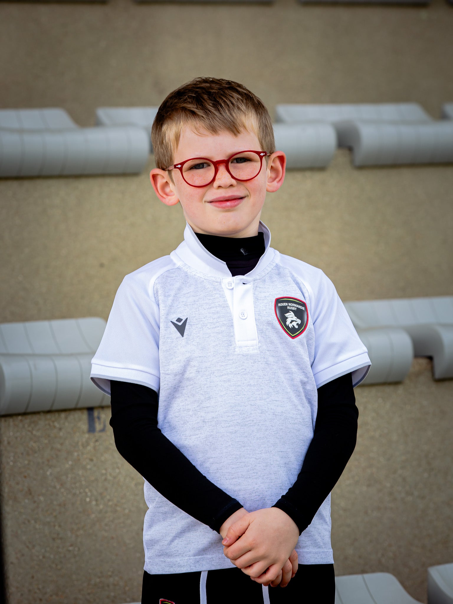 Un jeune garçon aux cheveux blonds courts et aux lunettes rouges sourit à la caméra devant les sièges vides du stade, portant un Polo Rugby Rouen Normandie Blanc Aulos JR par-dessus son maillot de corps noir.