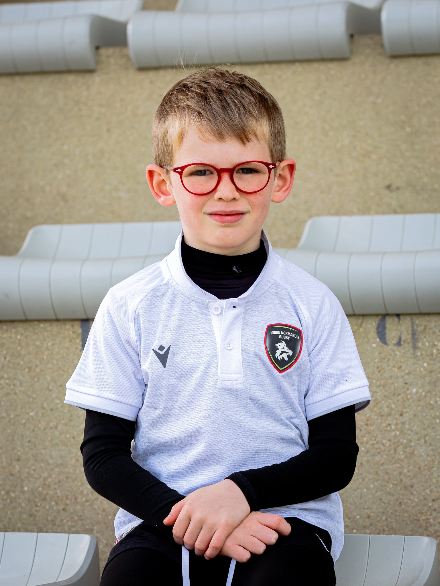 Un jeune garçon aux cheveux courts châtains clairs et aux lunettes rouges est assis sur les marches du stade. Il porte un Polo Rouen Normandie Rugby Blanc Aulos JR avec un maillot de corps noir et l'écusson du club sur la poitrine.