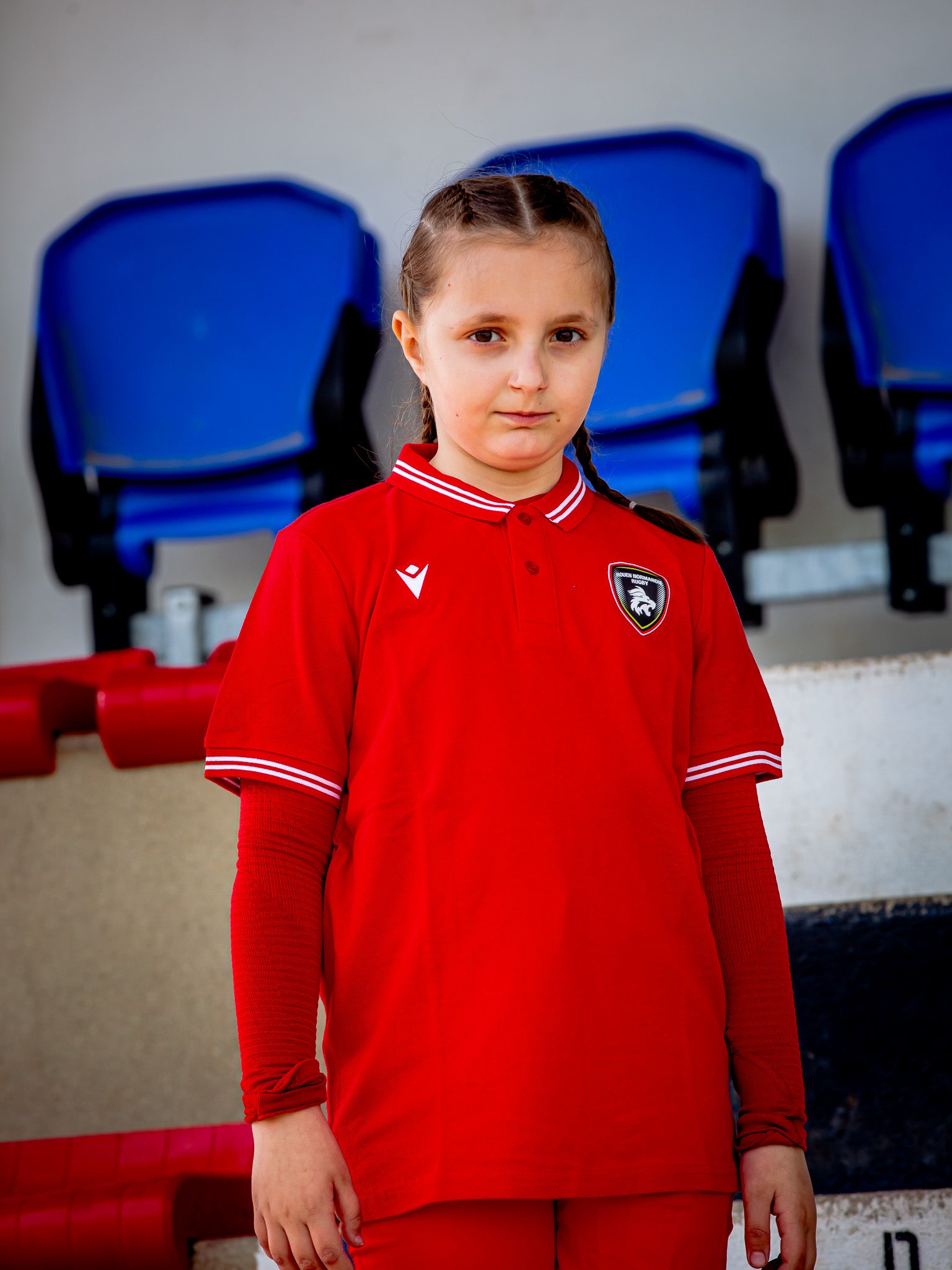 Une jeune fille aux cheveux tressés se tient devant les sièges bleus du stade, portant le maillot et le pantalon de supporter du Rouen Normandie Rugby Polo Yukar JR, regardant la caméra avec une expression neutre.