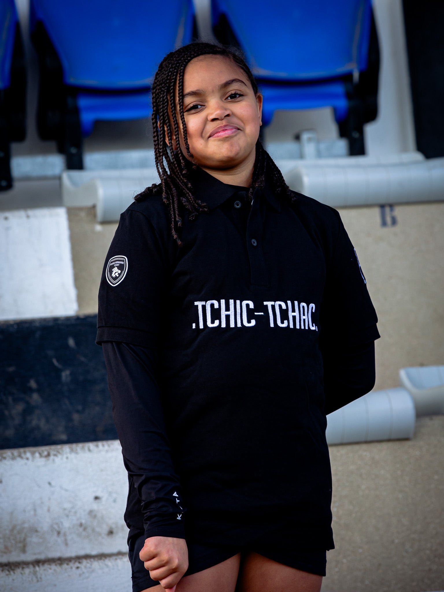 Une jeune fille souriante aux cheveux tressés porte le Polo manches courtes Tchic Tchac JR du Rouen Normandie Rugby, face à des sièges de stade bleus et un mur de béton.