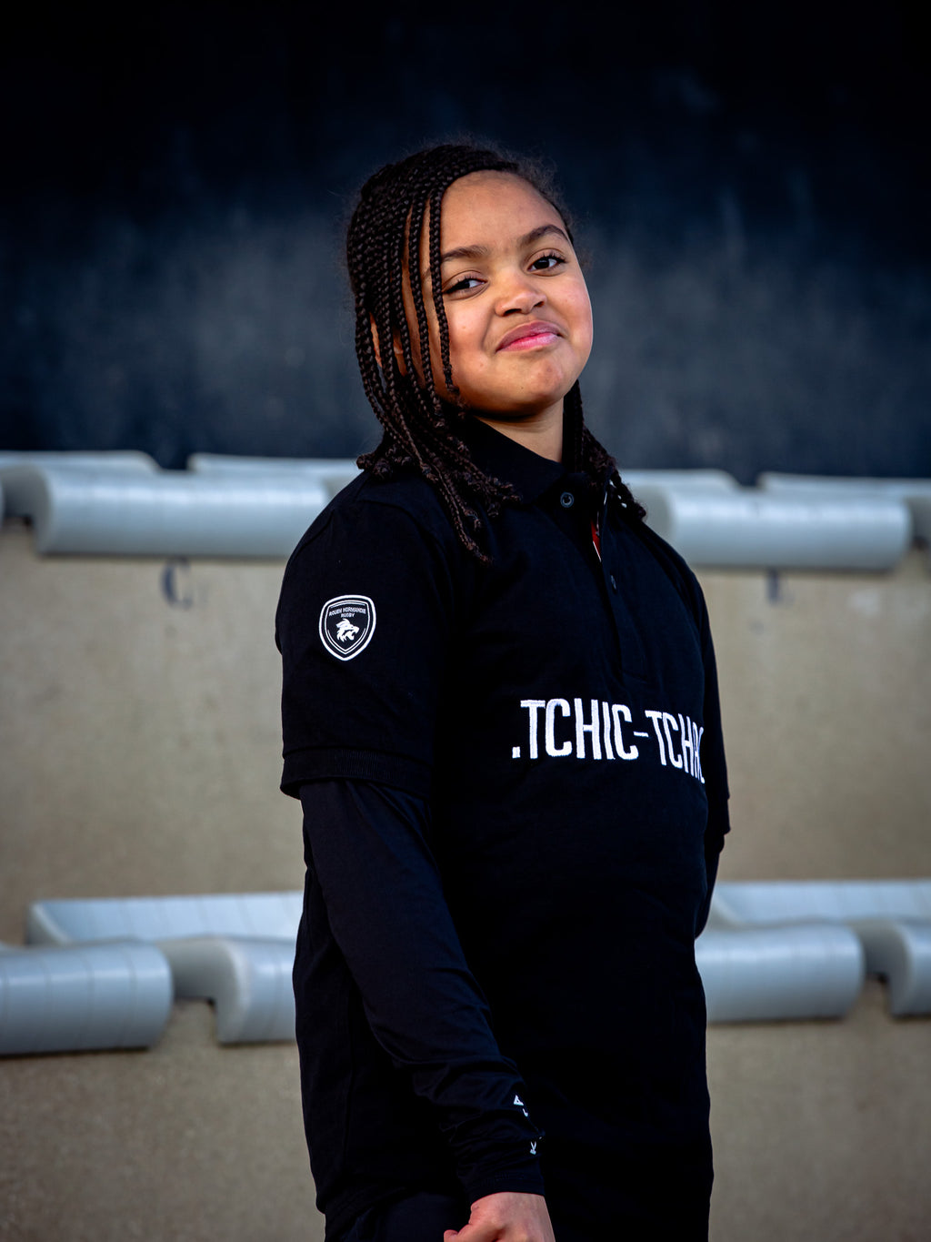 Une jeune fille aux cheveux tressés sourit légèrement devant les gradins du stade, portant le Polo Rouen Normandie Rugby manches courtes Tchic Tchac JR.