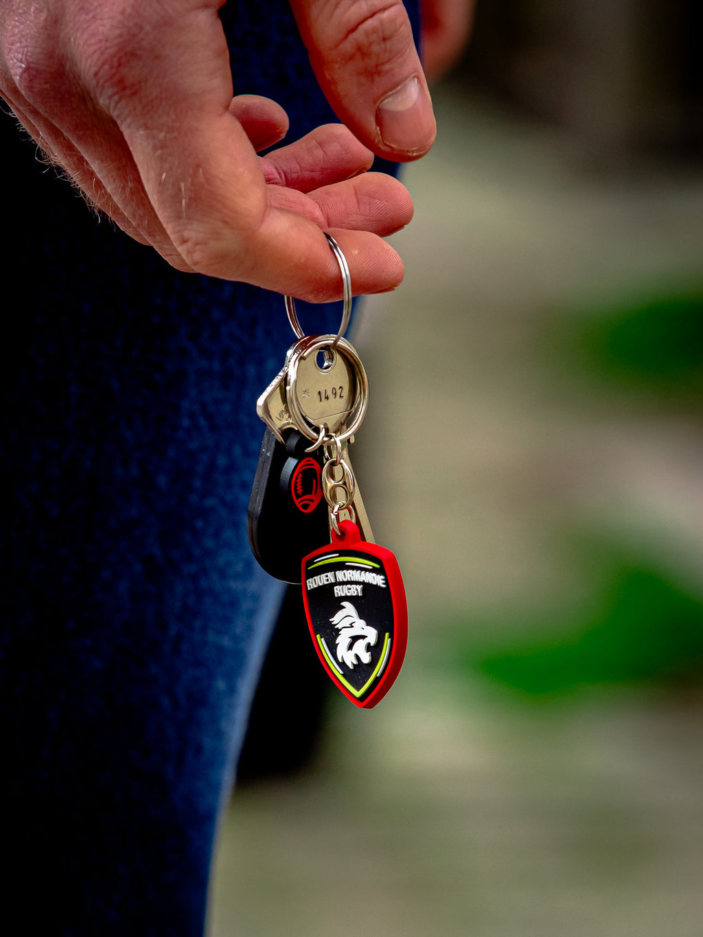 Une main tient un trousseau de clés avec un Porte-Clés RNR de Rouen Normandie Rugby, présentant un écusson rouge, noir et vert et un emblème blanc en forme de tête de lion, sur un fond flou.