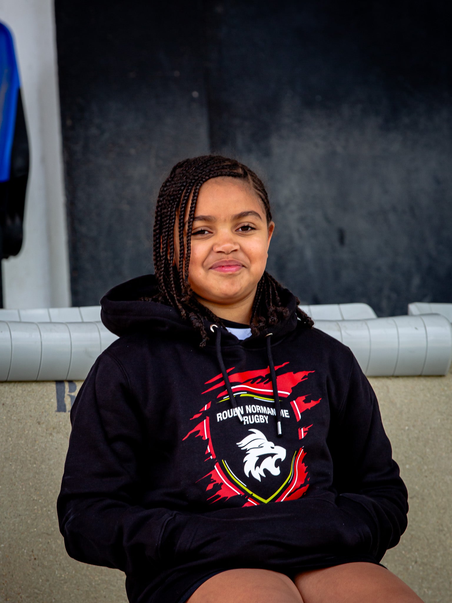 Une jeune fille aux cheveux tressés est assise et sourit dans le sweat à capuche Rouen Normandie Rugby Sweat Griffe JR, rayonnant de confort sur un fond neutre.