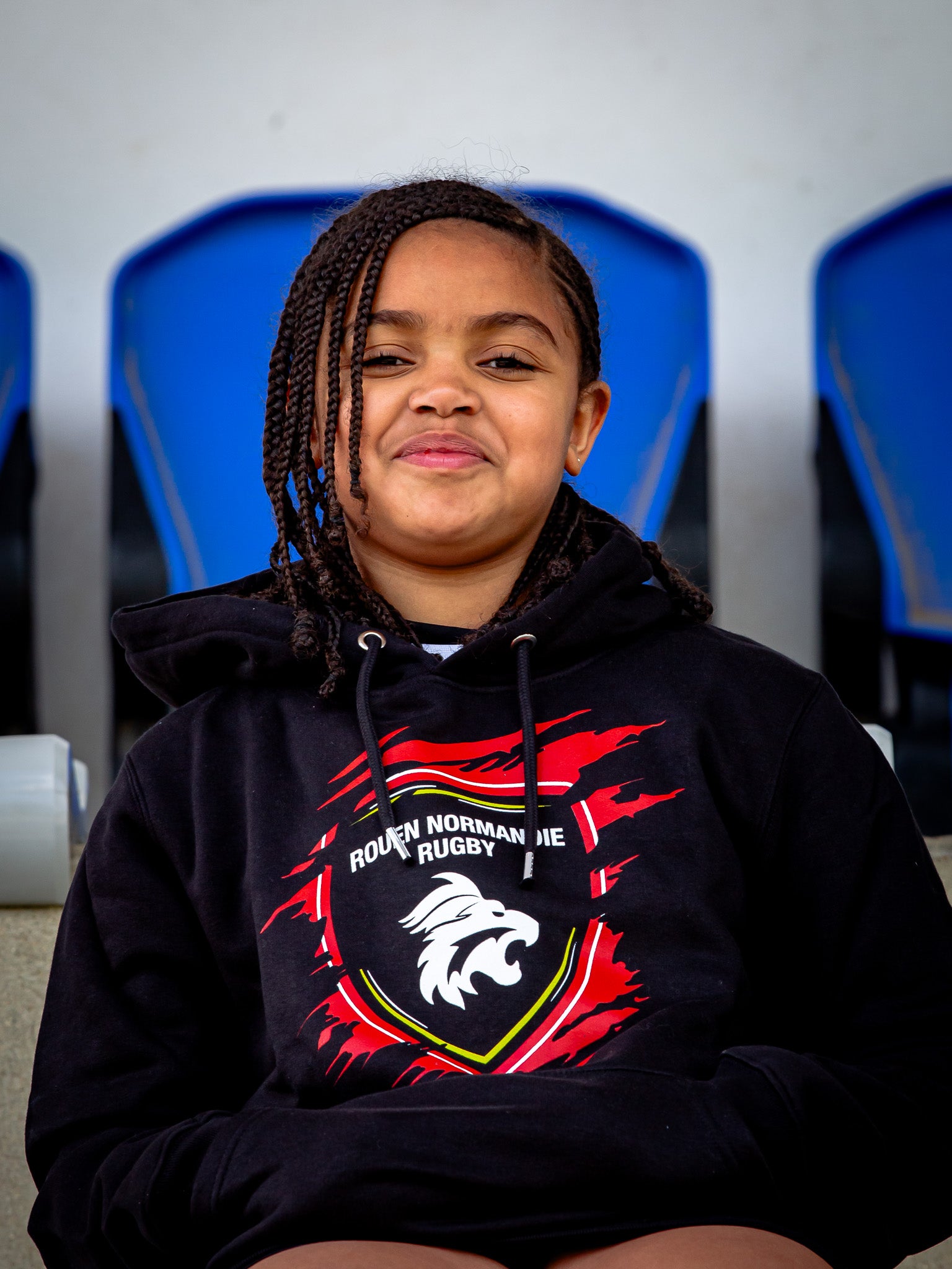 Une jeune fille aux cheveux tressés, rayonnante de confort, est assise, souriante, dans un Sweat Griffe JR du Rouen Normandie Rugby, dont les sièges bleus sont flous en arrière-plan.