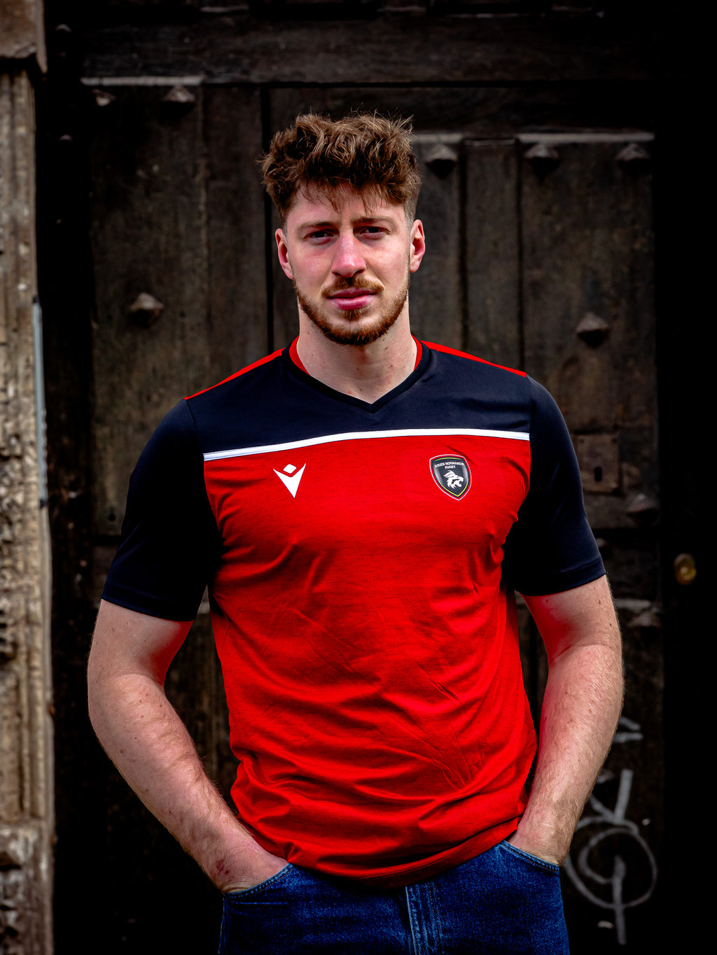 Un jeune homme aux cheveux bruns courts et à la barbe se tient devant une porte en bois sombre et rustique, portant le T-shirt Deneb Rouen Normandie Rugby - Macron by Rouen Normandie Rugby, les mains dans les poches de son jean.
