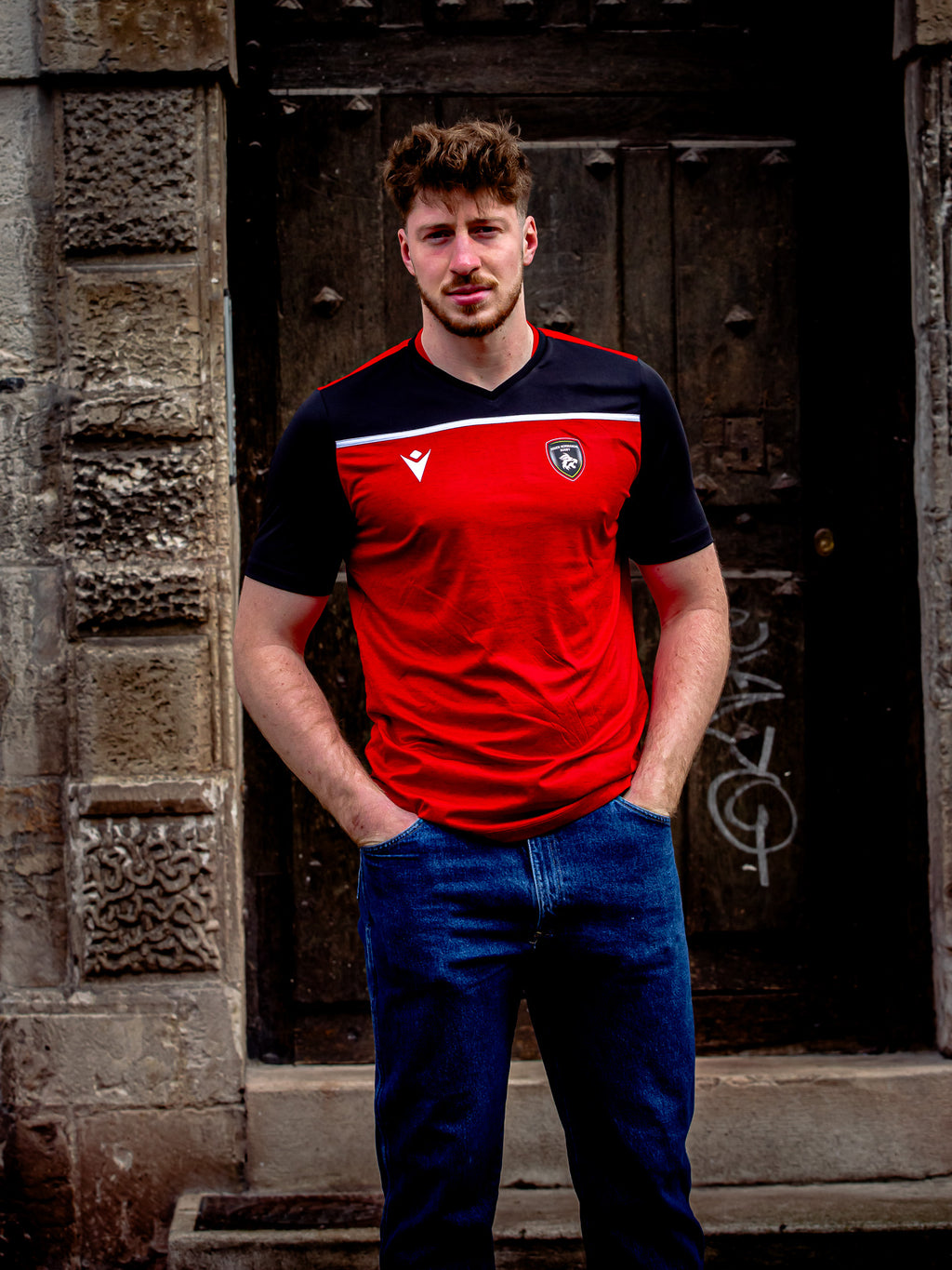 Un homme aux cheveux bruns courts se tient devant un mur de pierre usé par les intempéries et une vieille porte en bois, les mains dans les poches. Il porte un jean bleu et un T-shirt rouge et noir Deneb Rouen Normandie Rugby by Rouen Normandie Rugby.