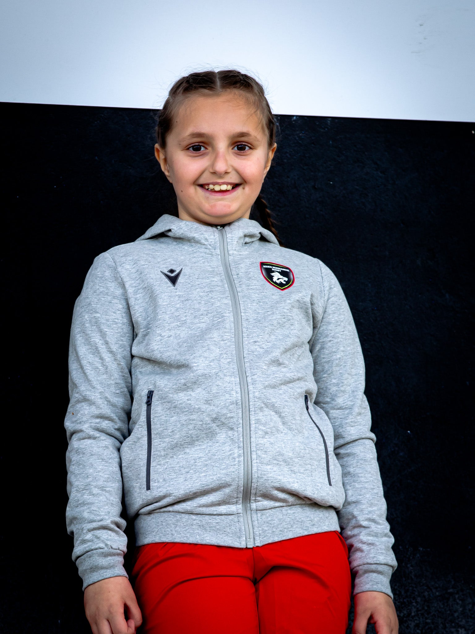 Une jeune fille aux cheveux tressés, souriante et portant la Veste Cello JR du Rouen Normandie Rugby avec un insigne sur la poitrine et un pantalon rouge vif, se tient devant un fond noir et blanc.