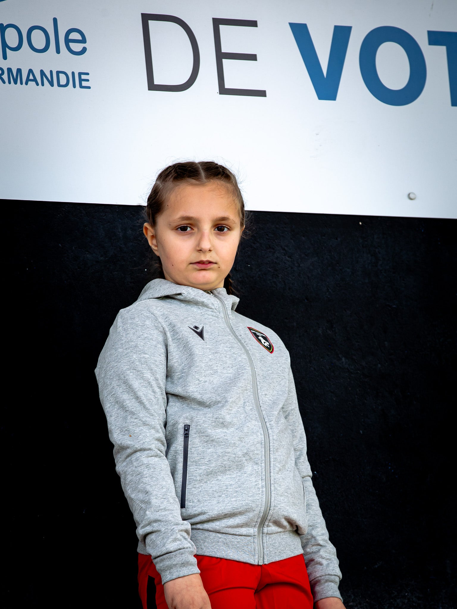 Une jeune fille aux cheveux tressés, vêtue d'une veste Cello JR de Rouen Normandie Rugby et d'un pantalon rouge, se tient devant un mur noir sur lequel est partiellement visible un panneau blanc avec des lettres noires et bleues en gras.